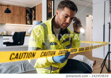 Police detective examining evidence with flashlight at a murder scene, caution tape visible in a modern apartment interior with wooden cabinets and furniture 142454639