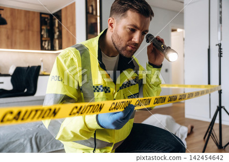 Police detective examining a crime scene with a flashlight while crouching near caution tape in a modern interior setting with a body outline visible on the floor Police detective examining a crime scene with a flashlight while crouching near caution tape in a modern interior setting with a body outline visible on the floor 142454642