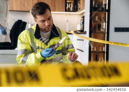 Forensic expert in yellow police jacket examines knife evidence at a murder scene, with caution tape and a modern kitchen visible in the background Forensic expert in yellow police jacket examines knife evidence at a murder scene, with caution tape and a modern kitchen visible in the background 142454658