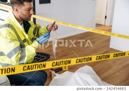Forensic expert in reflective police jacket examines evidence at a murder scene, with caution tape surrounding a covered body on a wooden floor in a residential setting Forensic expert in reflective police jacket examines evidence at a murder scene, with caution tape surrounding a covered body on a wooden floor in a residential setting 142454663