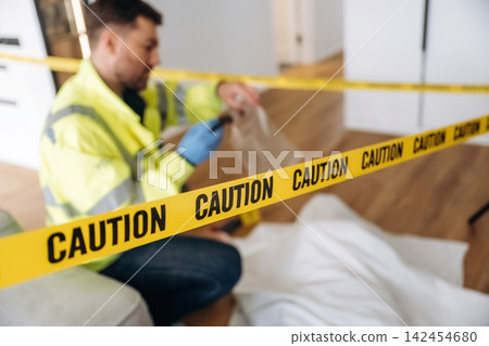 Forensic expert in yellow safety jacket collecting evidence at a murder scene, with caution tape and a covered body visible in a residential interior setting Forensic expert in yellow safety jacket collecting evidence at a murder scene, with caution tape and a covered body visible in a residential interior setting 142454680