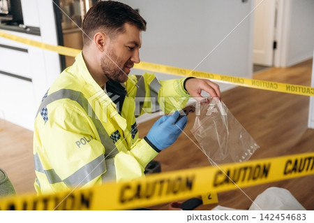Male forensic expert in yellow police jacket examines evidence inside a crime scene marked with caution tape, showcasing a modern interior setting Male forensic expert in yellow police jacket examines evidence inside a crime scene marked with caution tape, showcasing a modern interior setting 142454683