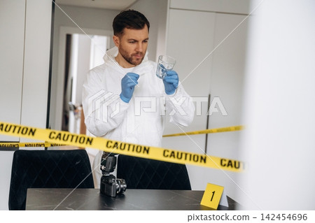 Forensic expert in white protective suit examines evidence in glass container at a crime scene, with caution tape and a camera on the table 142454696