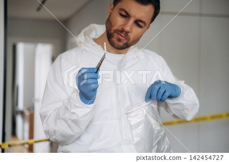 Forensic expert in white protective suit examines evidence with tweezers inside a crime scene marked with police tape, focused on collecting samples for investigation Forensic expert in white protective suit examines evidence with tweezers inside a crime scene marked with police tape, focused on collecting samples for investigation 142454727