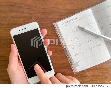 A woman's hand holding a smartphone and a November schedule book placed on a wooden table. A woman's hand holding a smartphone and a November schedule book placed on a wooden table. 142455257