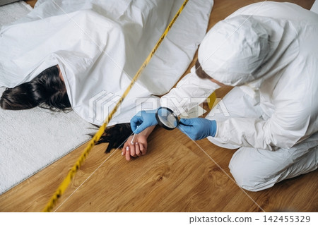 Forensic expert in protective suit examines evidence at a murder scene with a covered body on the floor and police tape marking the area Forensic expert in protective suit examines evidence at a murder scene with a covered body on the floor and police tape marking the area 142455329