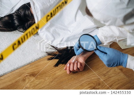 Forensic expert examining evidence at a murder scene with a magnifying glass, caution tape visible around the body on a wooden floor Forensic expert examining evidence at a murder scene with a magnifying glass, caution tape visible around the body on a wooden floor 142455330