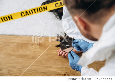 Forensic expert examining evidence at a murder scene, wearing gloves and protective clothing, with caution tape and a body visible in the background Forensic expert examining evidence at a murder scene, wearing gloves and protective clothing, with caution tape and a body visible in the background 142455333