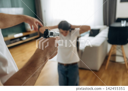 Armed assailant aims a pistol at a frightened person with hands raised inside a living room, conveying fear, danger, domestic threat, crime scene tension and urgent confrontation. Armed assailant aims a pistol at a frightened person with hands raised inside a living room, conveying fear, danger, domestic threat, crime scene tension and urgent confrontation. 142455351
