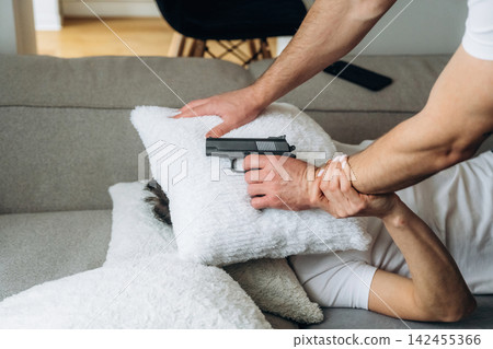 Male figure holding a handgun against a person's head while they lie on a couch covered with white pillows in a domestic interior setting Male figure holding a handgun against a person's head while they lie on a couch covered with white pillows in a domestic interior setting 142455366