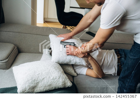 Male and female individuals playfully engage in a pillow fight on a gray sofa in a cozy living room with a green blanket and natural light streaming through the window Male and female individuals playfully engage in a pillow fight on a gray sofa in a cozy living room with a green blanket and natural light streaming through the window 142455367