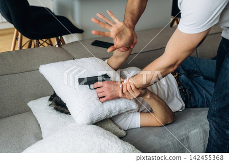 Male figure holding a handgun against a person's head while they are lying on a couch, surrounded by pillows in a domestic living room setting Male figure holding a handgun against a person's head while they are lying on a couch, surrounded by pillows in a domestic living room setting 142455368