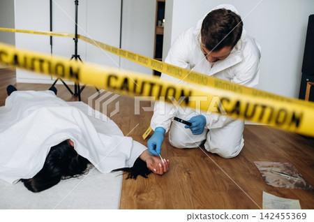 Forensic expert in white suit examines evidence at a murder scene with caution tape surrounding a body on the floor in a residential setting 142455369
