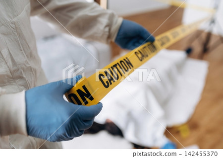 Forensic expert in protective gear holds yellow caution tape at a crime scene with covered evidence and wooden floor visible in the background Forensic expert in protective gear holds yellow caution tape at a crime scene with covered evidence and wooden floor visible in the background 142455370