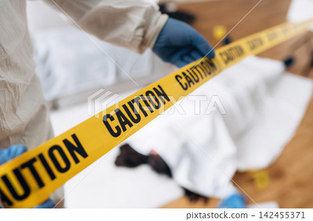 Forensic expert in protective suit holds yellow caution tape at a crime scene with a covered body and evidence markers visible on the floor Forensic expert in protective suit holds yellow caution tape at a crime scene with a covered body and evidence markers visible on the floor 142455371
