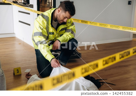 Police detective in reflective jacket examines evidence at a murder scene, with caution tape and a covered body visible on the wooden floor of a modern interior Police detective in reflective jacket examines evidence at a murder scene, with caution tape and a covered body visible on the wooden floor of a modern interior 142455382