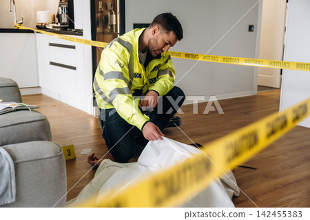 Police detective examining a body covered with a white sheet at a crime scene marked with caution tape in a modern interior setting with wooden flooring Police detective examining a body covered with a white sheet at a crime scene marked with caution tape in a modern interior setting with wooden flooring 142455383