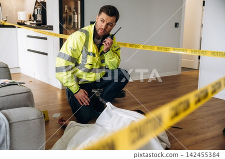 Male forensic expert in yellow safety jacket kneeling beside a covered body at a crime scene, police tape marking the area in a modern apartment interior Male forensic expert in yellow safety jacket kneeling beside a covered body at a crime scene, police tape marking the area in a modern apartment interior 142455384
