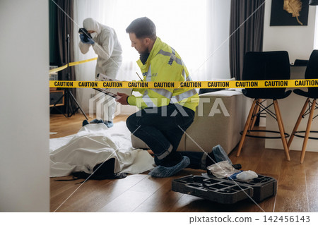 Forensic expert in yellow jacket examines evidence at a murder scene with caution tape, while police detective photographs the area in a residential setting Forensic expert in yellow jacket examines evidence at a murder scene with caution tape, while police detective photographs the area in a residential setting 142456143