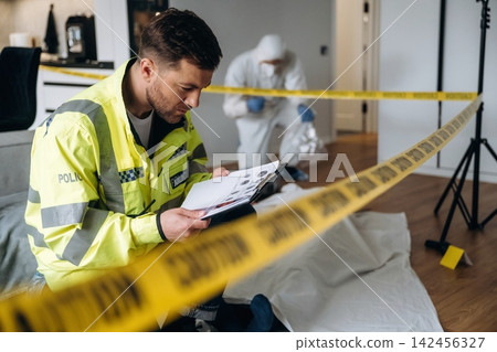 Police detective in yellow jacket examines evidence notebook at a murder scene, with forensic expert collecting evidence behind caution tape in a modern apartment Police detective in yellow jacket examines evidence notebook at a murder scene, with forensic expert collecting evidence behind caution tape in a modern apartment 142456327