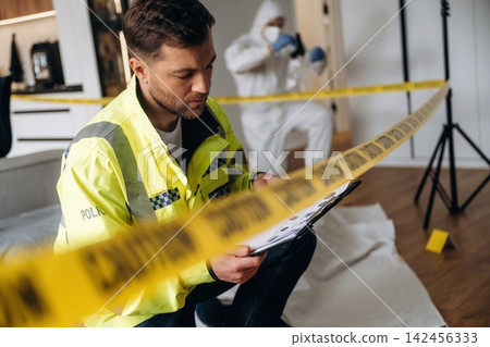 Police detective in yellow jacket examines evidence clipboard at a murder scene, with forensic expert in protective suit collecting samples in the background Police detective in yellow jacket examines evidence clipboard at a murder scene, with forensic expert in protective suit collecting samples in the background 142456333