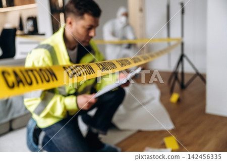 Forensic expert in yellow safety jacket kneeling near caution tape while examining evidence at a crime scene with a figure in protective suit in the background Forensic expert in yellow safety jacket kneeling near caution tape while examining evidence at a crime scene with a figure in protective suit in the background 142456335