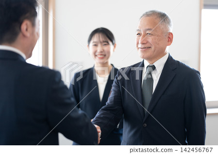 身著西裝的高級商務人士在辦公室裡互相微笑握手,女員工們在一旁觀看。 身著西裝的高級商務人士在辦公室裡互相微笑握手,女員工們在一旁觀看。 142456767