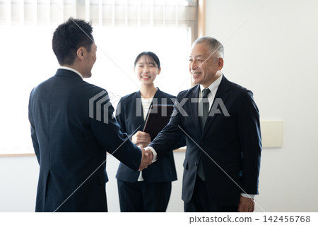 身著西裝的高級商務人士在辦公室裡互相微笑握手,女員工們在一旁觀看。 身著西裝的高級商務人士在辦公室裡互相微笑握手,女員工們在一旁觀看。 142456768
