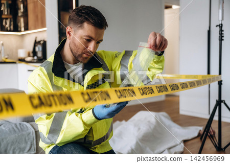 Forensic expert in yellow jacket examining evidence at a murder scene, with caution tape and a covered body visible in a modern interior setting Forensic expert in yellow jacket examining evidence at a murder scene, with caution tape and a covered body visible in a modern interior setting 142456969