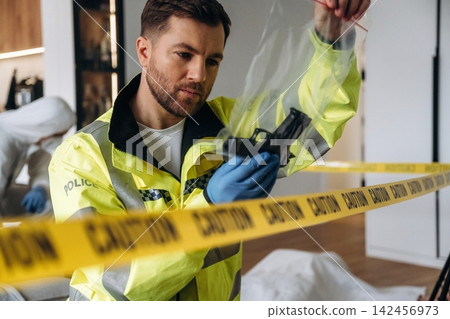 Male forensic expert in yellow police jacket examines firearm evidence inside a crime scene marked by caution tape and a covered body in a residential setting Male forensic expert in yellow police jacket examines firearm evidence inside a crime scene marked by caution tape and a covered body in a residential setting 142456973