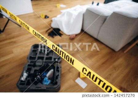 Forensic expert investigates a murder scene marked by caution tape, with a covered body and evidence markers on the wooden floor near a gray sofa Forensic expert investigates a murder scene marked by caution tape, with a covered body and evidence markers on the wooden floor near a gray sofa 142457270
