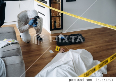Forensic expert in white protective suit photographing evidence at a murder scene with caution tape and a covered body on the floor in a modern interior setting Forensic expert in white protective suit photographing evidence at a murder scene with caution tape and a covered body on the floor in a modern interior setting 142457294