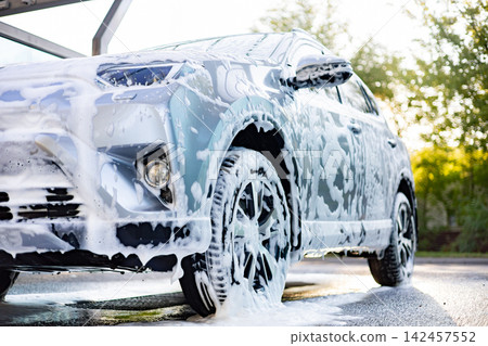 A silver SUV is covered in thick white foam during a thorough car wash process outdoors A silver SUV is covered in thick white foam during a thorough car wash process outdoors 142457552