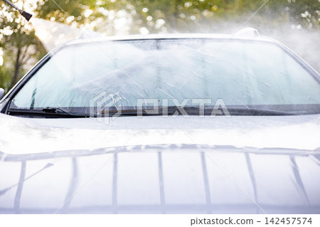 A person is using a pressure washer to clean the windshield of a white car outdoors A person is using a pressure washer to clean the windshield of a white car outdoors 142457574