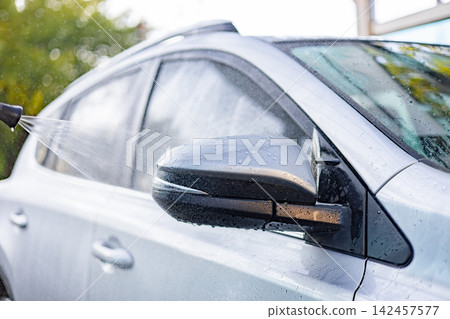A person is washing a car with a hose, spraying water onto the side mirror and bodywork A person is washing a car with a hose, spraying water onto the side mirror and bodywork 142457577