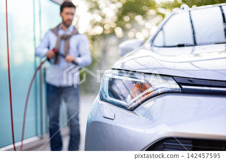 A caucasian man is using a high-pressure washer to clean his silver car outdoors on a sunny day A caucasian man is using a high-pressure washer to clean his silver car outdoors on a sunny day 142457595