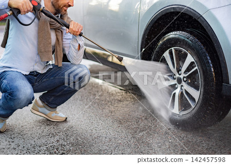 A caucasian man wearing jeans and a light blue shirt is squatting down to pressure wash the wheel of a silver car A caucasian man wearing jeans and a light blue shirt is squatting down to pressure wash the wheel of a silver car 142457598