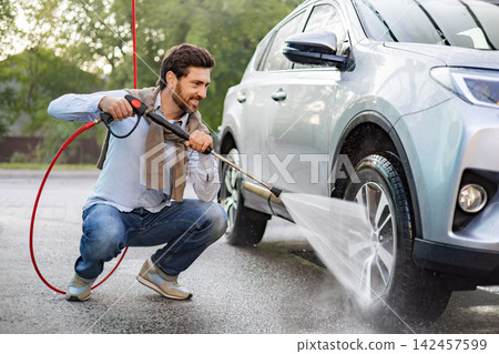 A smiling Caucasian man is crouching down to wash his car using a high-pressure water jet on the wheel and lower body of the vehicle A smiling Caucasian man is crouching down to wash his car using a high-pressure water jet on the wheel and lower body of the vehicle 142457599