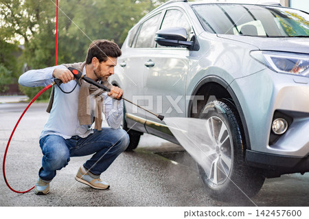 A caucasian man is kneeling down and using a high-pressure washer to clean the wheels and lower body of his silver SUV 142457600