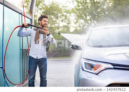 A man is using a high-pressure water jet to clean his car at an outdoor self-service car wash station A man is using a high-pressure water jet to clean his car at an outdoor self-service car wash station 142457612