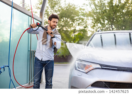 A man is actively cleaning his car using a high-pressure water spray at an outdoor self-service car wash station A man is actively cleaning his car using a high-pressure water spray at an outdoor self-service car wash station 142457625