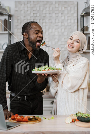A man and woman of diverse backgrounds share a moment of joy while preparing and tasting a fresh salad in a bright kitchen setting A man and woman of diverse backgrounds share a moment of joy while preparing and tasting a fresh salad in a bright kitchen setting 142457635