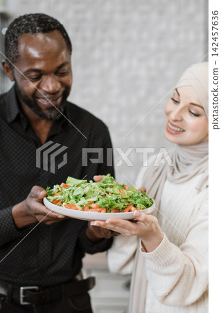 A smiling man and woman, one wearing a hijab, share a plate of fresh salad, representing healthy eating and togetherness A smiling man and woman, one wearing a hijab, share a plate of fresh salad, representing healthy eating and togetherness 142457636