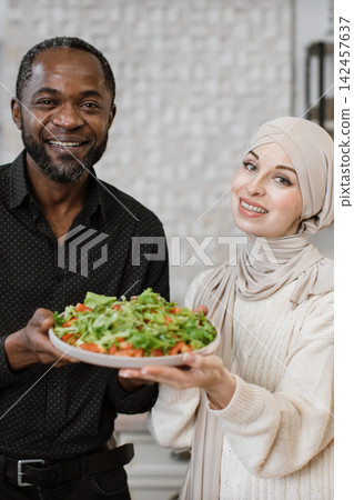 A smiling African man and a woman in a hijab present a large, fresh salad together, symbolizing healthy eating and partnership 142457637