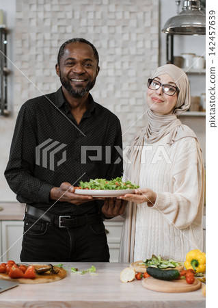 A smiling African man and a woman in hijab stand in a kitchen, holding a plate of fresh salad, surrounded by ingredients for a healthy meal A smiling African man and a woman in hijab stand in a kitchen, holding a plate of fresh salad, surrounded by ingredients for a healthy meal 142457639