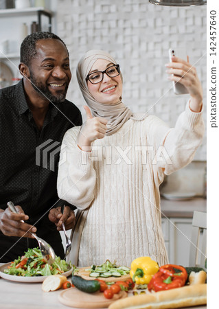 A smiling couple, one wearing a hijab, takes a selfie while preparing a healthy meal together in a bright kitchen A smiling couple, one wearing a hijab, takes a selfie while preparing a healthy meal together in a bright kitchen 142457640
