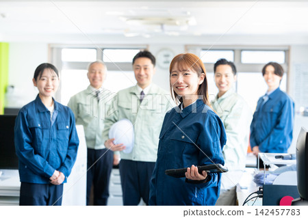 A woman in work clothes and staff members are smiling and looking at a tablet in the office. A woman in work clothes and staff members are smiling and looking at a tablet in the office. 142457783