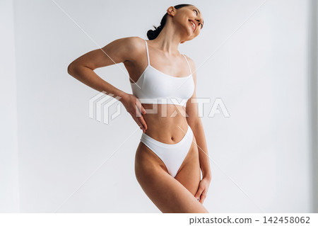 Slim female model in white lingerie poses gracefully against a plain background, showcasing a toned physique and confident posture in a fashion photography setting Slim female model in white lingerie poses gracefully against a plain background, showcasing a toned physique and confident posture in a fashion photography setting 142458062