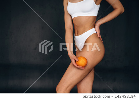 Slim female model in white bikini holding an orange against her thigh, showcasing her curves with a dark background emphasizing her figure and the fruit Slim female model in white bikini holding an orange against her thigh, showcasing her curves with a dark background emphasizing her figure and the fruit 142458184