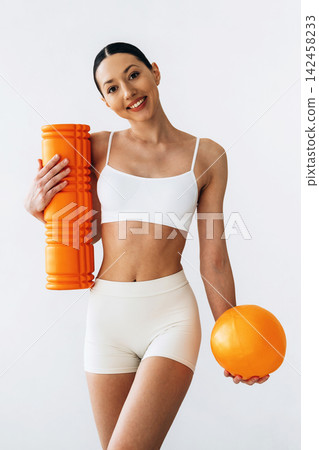 Slim female fitness model in white sportswear holding orange foam roller and ball, standing against a plain white background, showcasing athletic physique and workout gear Slim female fitness model in white sportswear holding orange foam roller and ball, standing against a plain white background, showcasing athletic physique and workout gear 142458233
